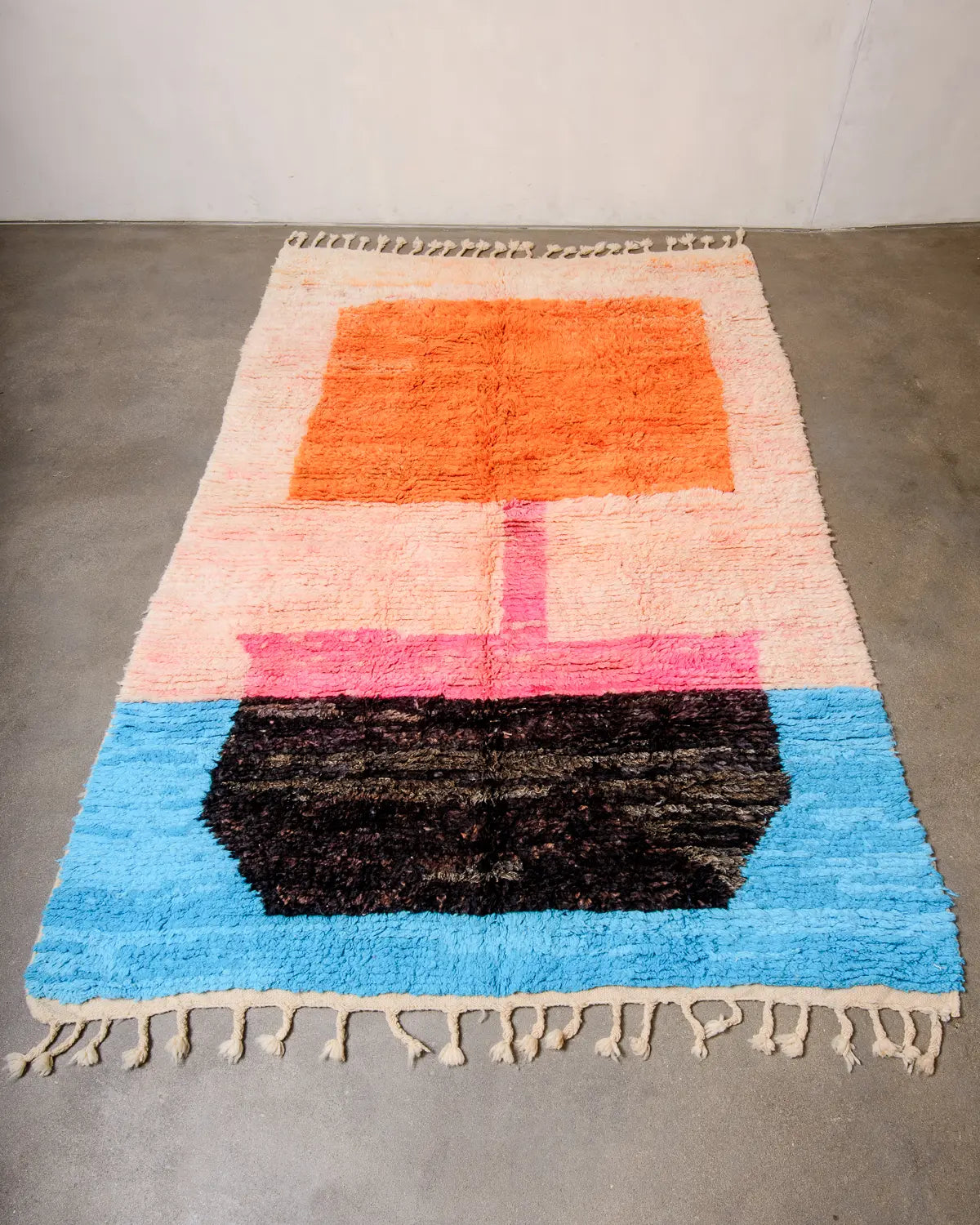 Wool Berber rug from Morocco in shades of peach, orange, pink, black and blue laying flat on concrete studio floor.